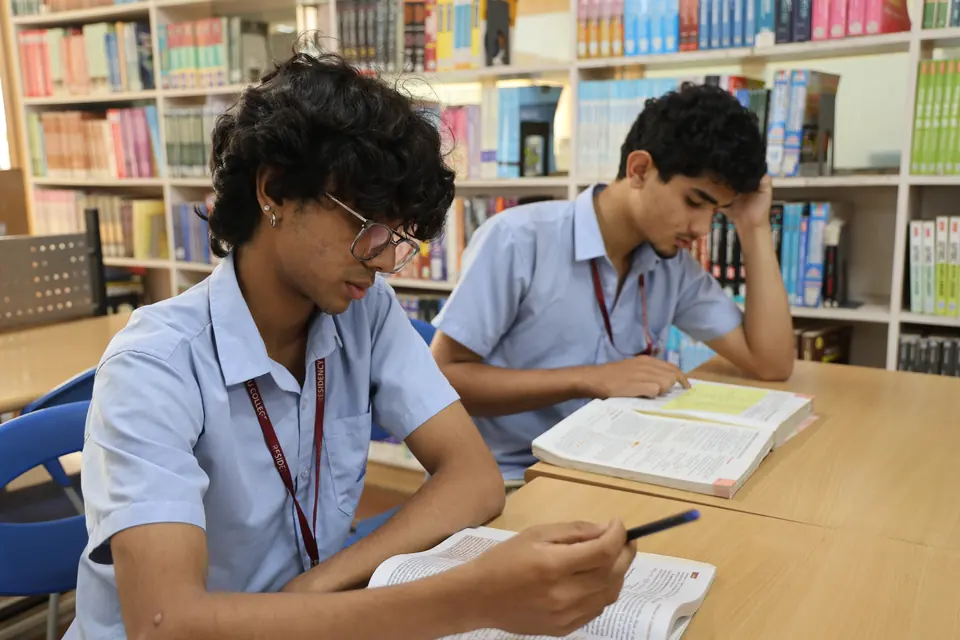 Two students reading at a library table in front of the bookshelves.