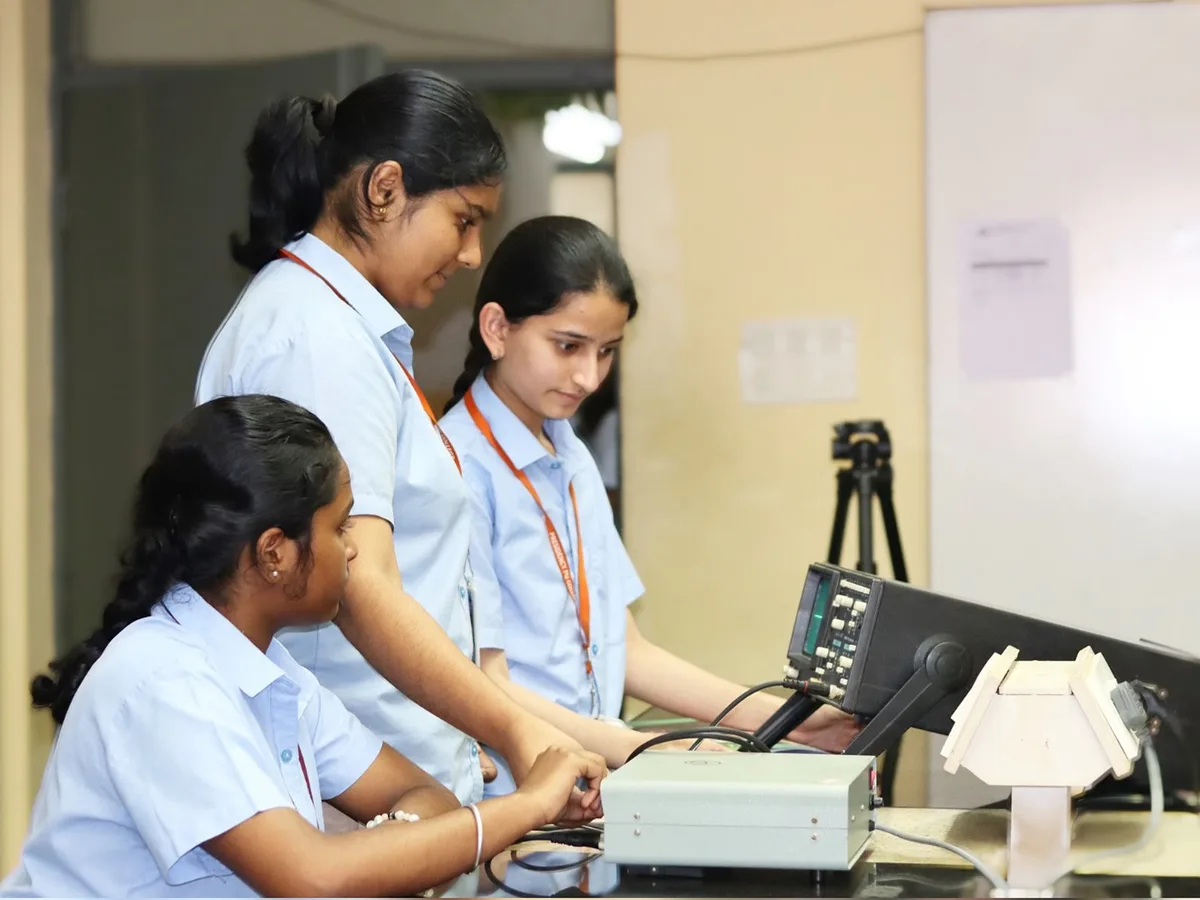 Students at a Physics lab bench, one adjusting an electronic instrument beside a tripod.